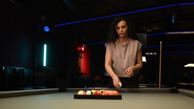 Focused woman arranging billiard balls on a pool table before starting a game in a dark club with neon lights