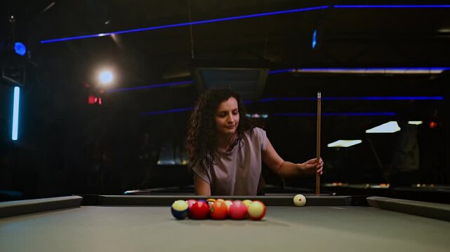 Focused curly haired woman breaking the rack while playing a game of pool in a dark, atmospheric billiard hall