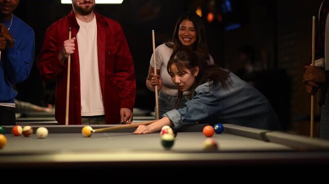 Young asian woman making a successful shot while playing pool. Her happy friends are watching, smiling and cheering for her victory