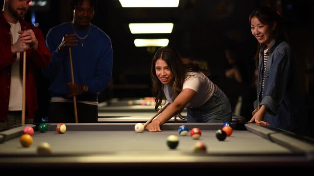 Cheerful young woman making a winning billiards shot while playing pool with multicultural friends in a dark bar