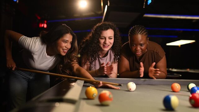 Three young multiracial women enjoying a night out, playing pool together. An asian woman makes a successful shot, and they celebrate happily