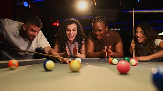 Multi ethnic group of friends having fun and playing billiards. Young man making a successful shot while his female friends cheer him on