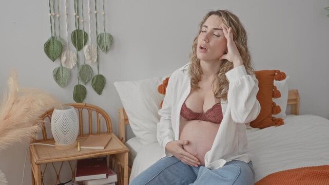 Pregnant woman sitting on bed in bedroom in open shirt and bra, cradling bare belly with one hand and hand on forehead for headache beside rattan nightstand; discomfort.