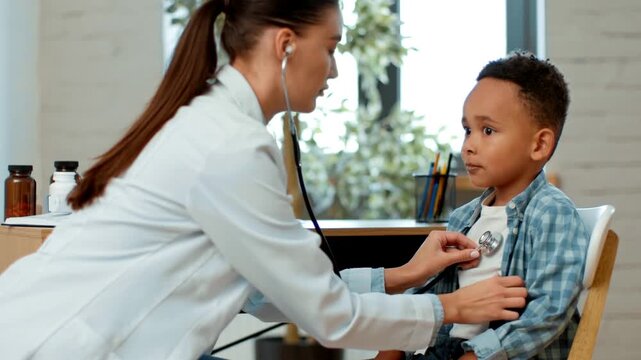A woman in a white coat uses a stethoscope to check the heart of a boy sitting in a chair. They are in a well-lit clinic with plants in the background. The boy looks attentive.