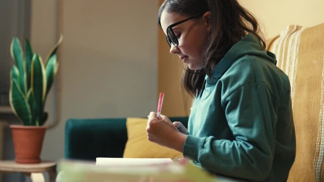 Smart Little Girl in Big Glasses Writing in Notebook while Sitting on Couch with Stack of Books &mdash; Cinematic 4K 10-bit