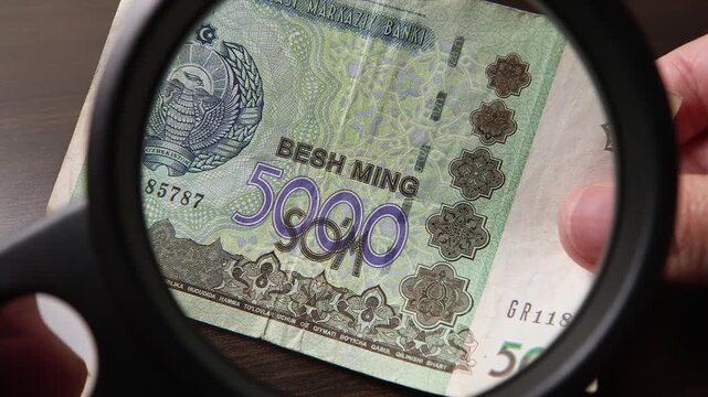 Examination of 5000 Uzbekistan Sum (UZS) cash money with magnfication glass, loupe. Concept of authenticity check, fight against counterfeiting of banknotes, anti fraud measures in Uzbekistan.