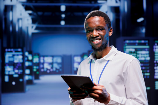 Portrait of smiling technician in front of supercomputers in high performance computing server room, ensuring flawless operations conditions. Expert monitoring data center hardware energy consumption