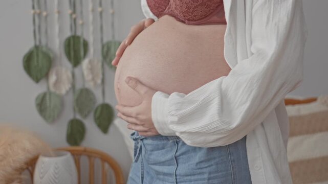 Pregnant woman cradling bare belly with both hands in studio near macrame wall and rattan chair, wearing open shirt and lace bra; serene anticipation.