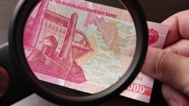 Examination of 2000 Uzbekistan Sum (UZS) cash money with magnfication glass, loupe. Concept of authenticity check, fight against counterfeiting of banknotes, anti fraud measures in Uzbekistan.