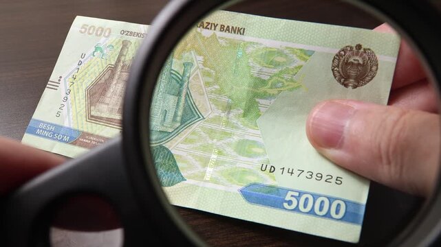 Examination of 5000 Uzbekistan Sum (UZS) cash money with magnfication glass, loupe. Concept of authenticity check, fight against counterfeiting of banknotes, anti fraud measures in Uzbekistan.