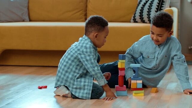 Two boys are sitting on the floor in a living room. One boy is stacking colorful blocks while the other observes. They are enjoying their time together in a cozy space.