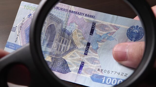 Examination of 10000 Uzbekistan Sum (UZS) cash money with magnfication glass, loupe. Concept of authenticity check, fight against counterfeiting of banknotes, anti fraud measures in Uzbekistan.