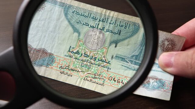 Examination of UAE Dirham (AED) cash money with magnfication glass, loupe. Concept of authenticity check, fight against counterfeiting of banknotes, anti fraud measures in UAE. Close up view.