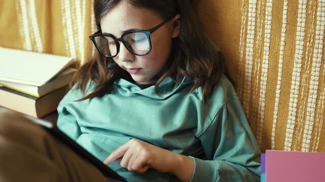 Little Girl in Glasses Using Digital Tablet on Couch with Stack of Books, Child Studying with Pad at Home, 4K 10-bit