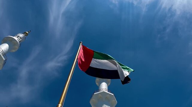 Low angle view of the United Arab Emirates flag waving on a flagpole against a clear blue sky with soft wispy clouds