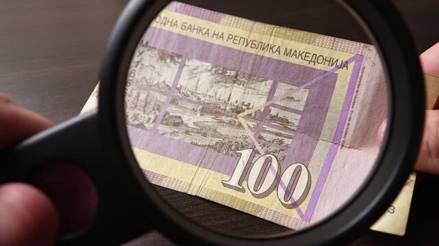 Examination of 100 Macedonian denar cash money with magnfication glass, loupe. Concept of authenticity check, fight against counterfeiting of banknotes, anti fraud measures in North Macedonia.