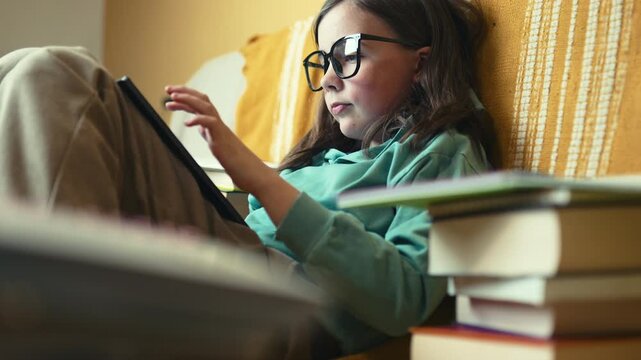 Little Girl in Glasses Using Digital Tablet on Couch with Stack of Books, Child Studying with Pad at Home, 4K 10-bit
