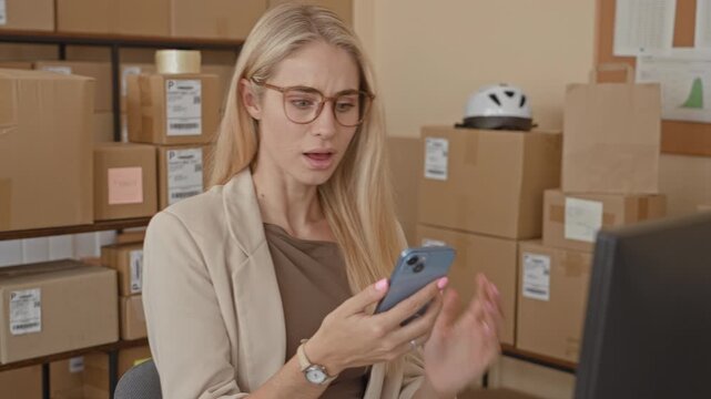 Woman holding smartphone with hand on cheek, looking at computer screen amid stacked parcels in a storage building; surprise.