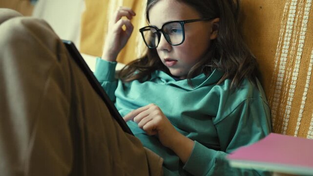 Little Girl in Glasses Using Digital Tablet on Couch with Stack of Books, Child Studying with Pad at Home, 4K 10-bit