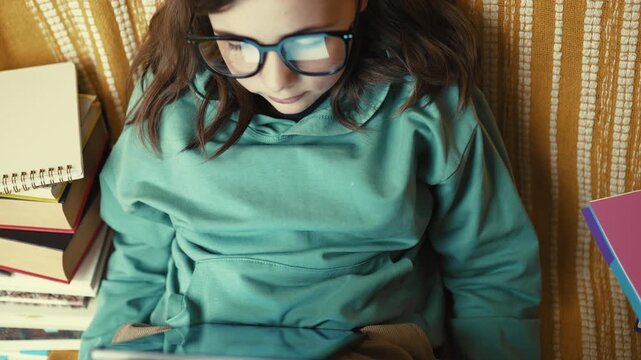 Little Girl in Glasses Using Digital Tablet on Couch with Stack of Books, Child Studying with Pad at Home, 4K 10-bit
