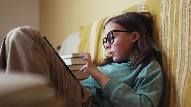 Little Girl in Glasses Using Digital Tablet on Couch with Stack of Books, Child Studying with Pad at Home, 4K 10-bit