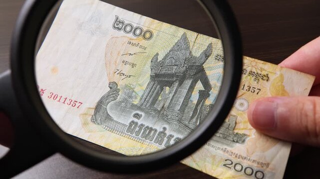 Examination of 2000 Cambodian Riel (KHR) cash money with magnfication glass, loupe. Concept of authenticity check, fight against counterfeiting of banknotes, anti fraud measures in Cambodia.