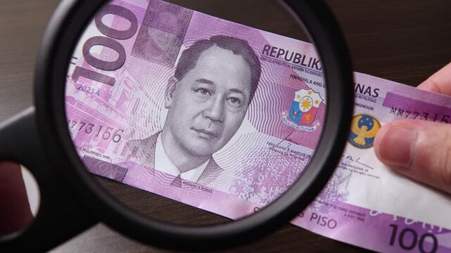 Examination of 100 Philippine peso (PHP) cash money with magnfication glass, loupe. Concept of authenticity check, fight against counterfeiting of banknotes, anti fraud measures in Philippines.