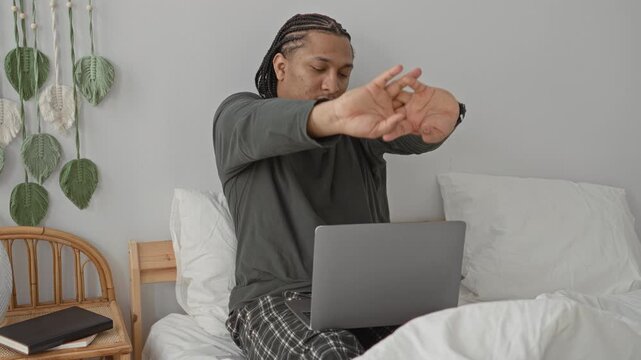 Man on bed with laptop, hands behind neck stretching while seated in a building bedroom, braided hair visible and pajama pants; calm concentration.