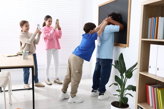 Bullying. Children filming boy pressing his African American classmate against chalkboard in classroom