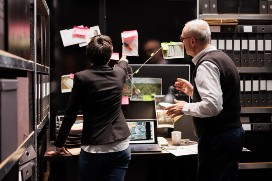 Private detectives looking at investigation board, analyzing crime scene evidence in arhive room. Elderly police officer explaining missing person report to investigator, working at criminal case