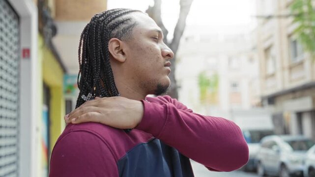 Man wearing maroon sweatshirt holding his shoulder with hand and gazing sideways on a city street; thoughtful introspection.