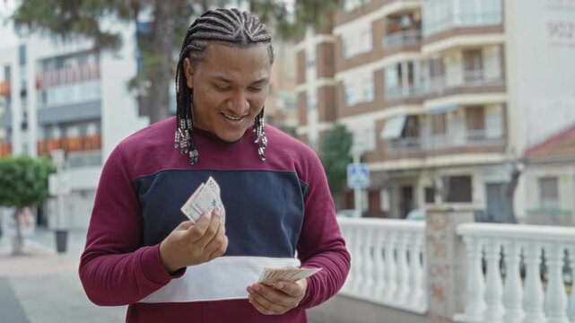 Young man counting pesos banknotes with both hands, smiling and looking down beside a white balustrade and apartment buildings on a city street; calm confidence.