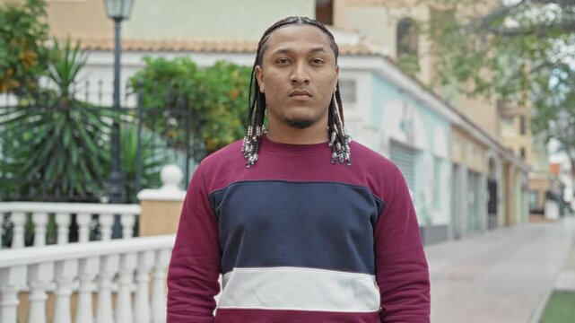 Man with braided hair and visible face standing and looking at camera on street with balustrade and shops; urban confident.