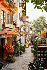 Fototapeta premium a quiet, charming alleyway features a narrow, stone-paved path flanked by colorful buildings, including one with a striking orange and yellow facade adorned with potted plants and traditional decorati