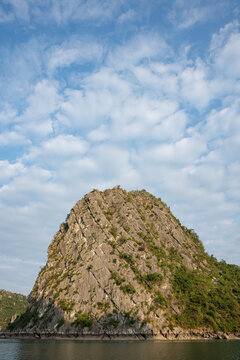 a dramatic limestone karst island rising steeply from calm waters under a bright, cloud-filled sky