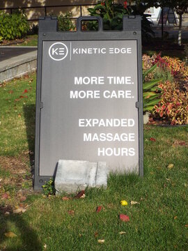 Ottawa, Ontario, Canada &ndash; August 02, 2025: A black sandwich board sign for "Kinetic Edge" sits on a lawn, advertising expanded massage hours and personalized care on Carling Ave.
