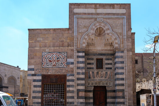 View of the Zawya and Sabil of Sultan Faraj Ibn Barquq featuring intricate stone carvings, ablaq striped masonry, and a decorative portal under a clear blue sky in Cairo, Egypt.
