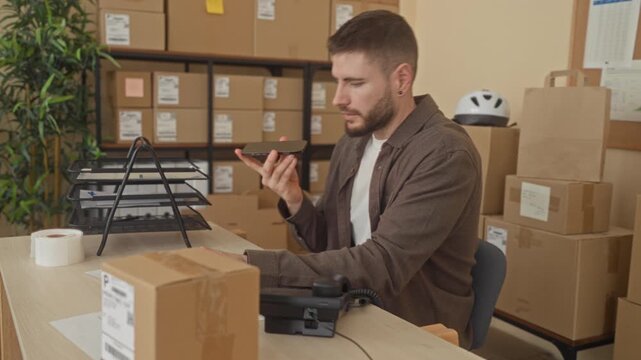 Man holding smartphone and checking parcel at a shipping desk inside a building surrounded by stacked boxes and packing tape; efficiency focus productivity.
