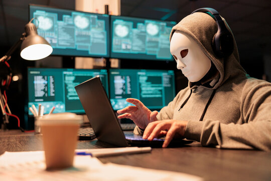 Hacker with mask and hood breaking security server, installing virus to create computer malware and steal big online data. Masked criminal scammer hacking netowrk system at night.