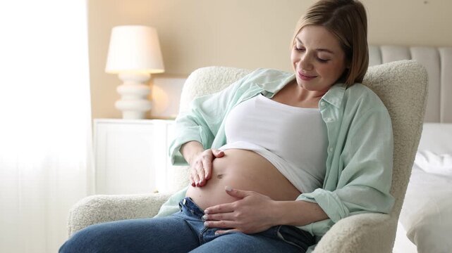 Pregnant woman touching her belly in armchair indoors, camera moving in
