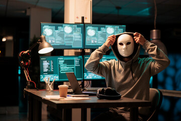 Scary masked hacker installing virus to hack system on computer, working late at night in office. Cyber terrorist and criminal with mask hacking network server and doing scam or fraud. © DC Studio