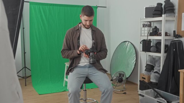 Young man adjusts camera with hands in studio green screen backdrop, sits on stool and smiles while checking settings; creative satisfaction.