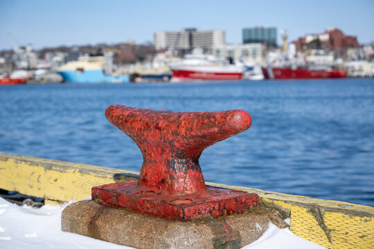A large red, rusty bollard on a wharf overlooking downtown St. John's, Newfoundland. There's a row of fishing boats and supply vessels in the background, moored in the harbour. It's a sunny day.