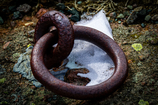 A vintage round cast-iron karabin climbing rappel metal ring embedded into a rocky cliff face. The bolted ring has snow under the loop. The corroded anchor bolt is thick and smaller than the ring.