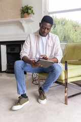 Obraz premium African American man sitting in living room on green armchair using tablet and tapping screen
