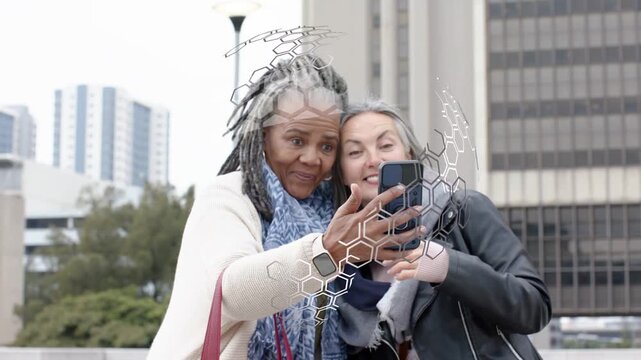 Left woman raising smartphone, starting hex overlay around faces and both posing for tech in plaza