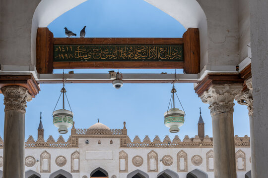 View of the historic Al-Azhar Mosque with its ornate white arches, hanging lanterns, and pigeons perched on a wooden beam under a clear blue sky Cairo, Egypt.