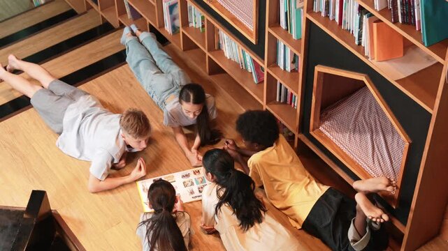 Group of diverse children lying down in circle while reading a book at library. Top view of girl sharing a magazine while pointing at interested topic and talking with lovely friends. Edification