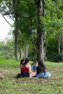 Two Latina mothers talking while children sit on grass during relaxed picnic in green park