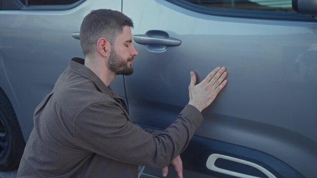 Young hispanic man kneels and touches a dent on a gray car door with bare hand while inspecting panel on street; concern.